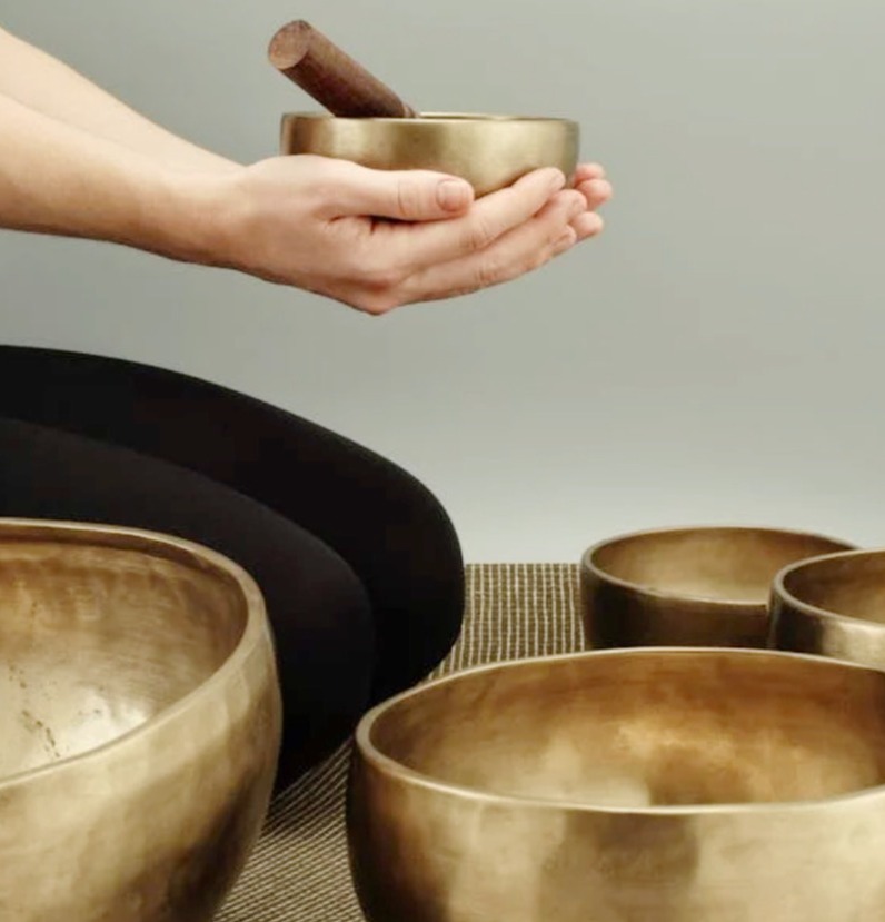 Depict a close-up of a singing bowl being played, with ripples of sound waves emanating from it. The background is blurred and suggests a dimly lit, peaceful space. Use warm, golden lighting to create a sense of tranquility.